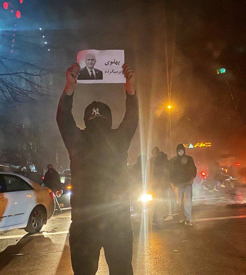 Masked demonstrator holding a portrait of Reza Pahlavi and a Persian sign
