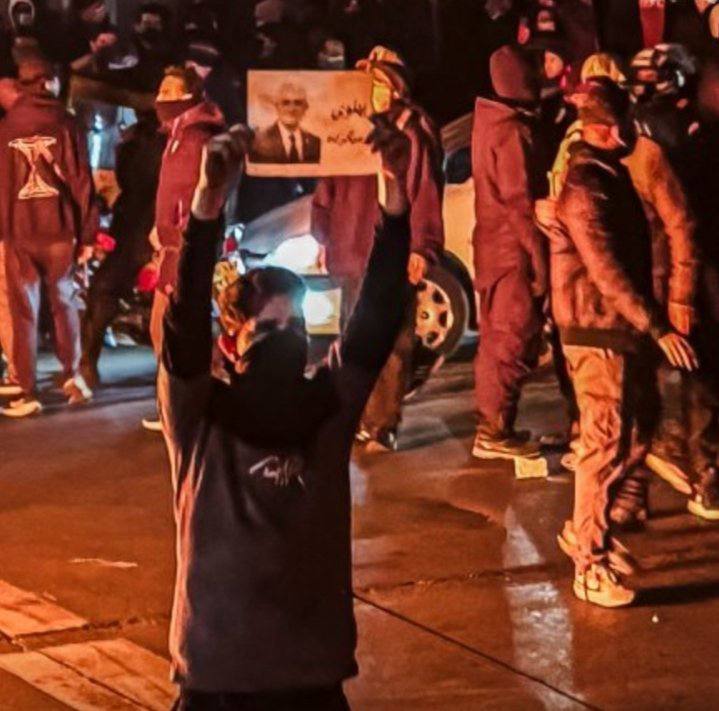 Masked demonstrator holding a portrait of Reza Pahlavi in a crowd